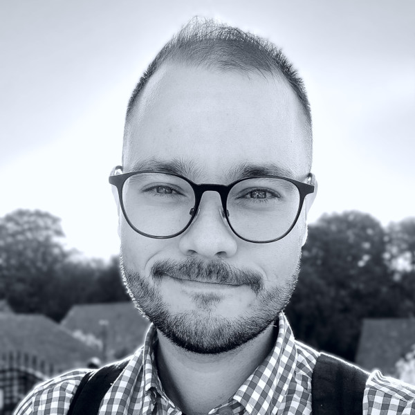 Monochromatická fotografia Martina Koleniča. Je usmiaty, má tmavé vlasy, strnisko a&nbsp;okuliare, na sebe má košeľu. v&nbsp;pozadí je rozmazaný strom.
